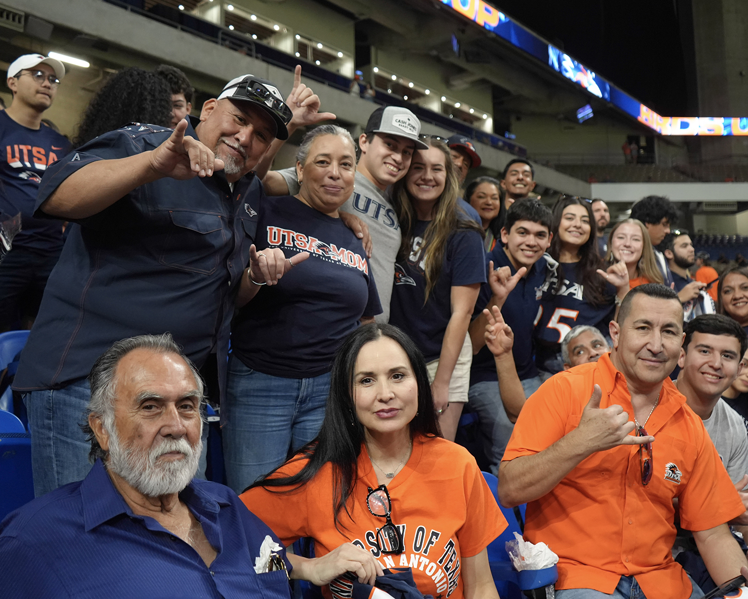 Multiple families in the stands at a game wearing blue and orange school shirts posing for a photo while holding the schools "birds up" hand gesture.